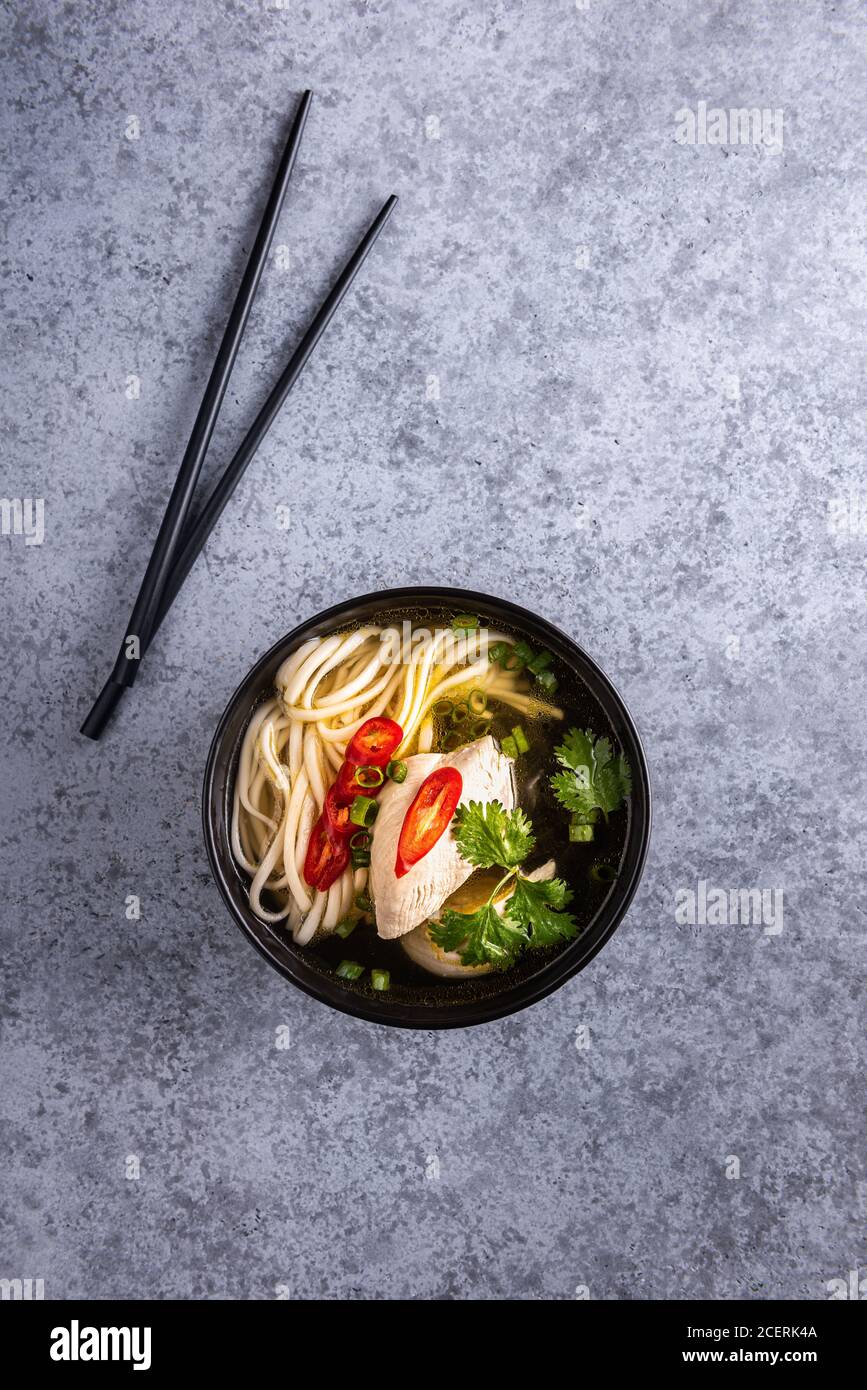 Würzige asiatische Suppe. Hühnerbouillon und Fleisch, rote Chilischote, frischer Koriander und Frühlingszwiebeln, japanische Nudeln in schwarzer Suppenschüssel mit Essstäbchen auf g Stockfoto