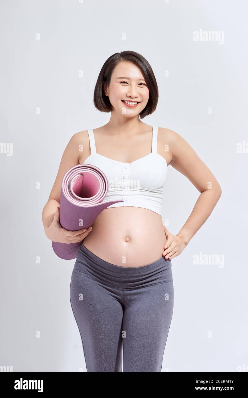 Junge attraktive schwangere Frau berühren ihren Bauch, während Yoga-Matte in den Händen halten, schwangere Mutter tun körperliche Übungen, posiert isoliert über w Stockfoto