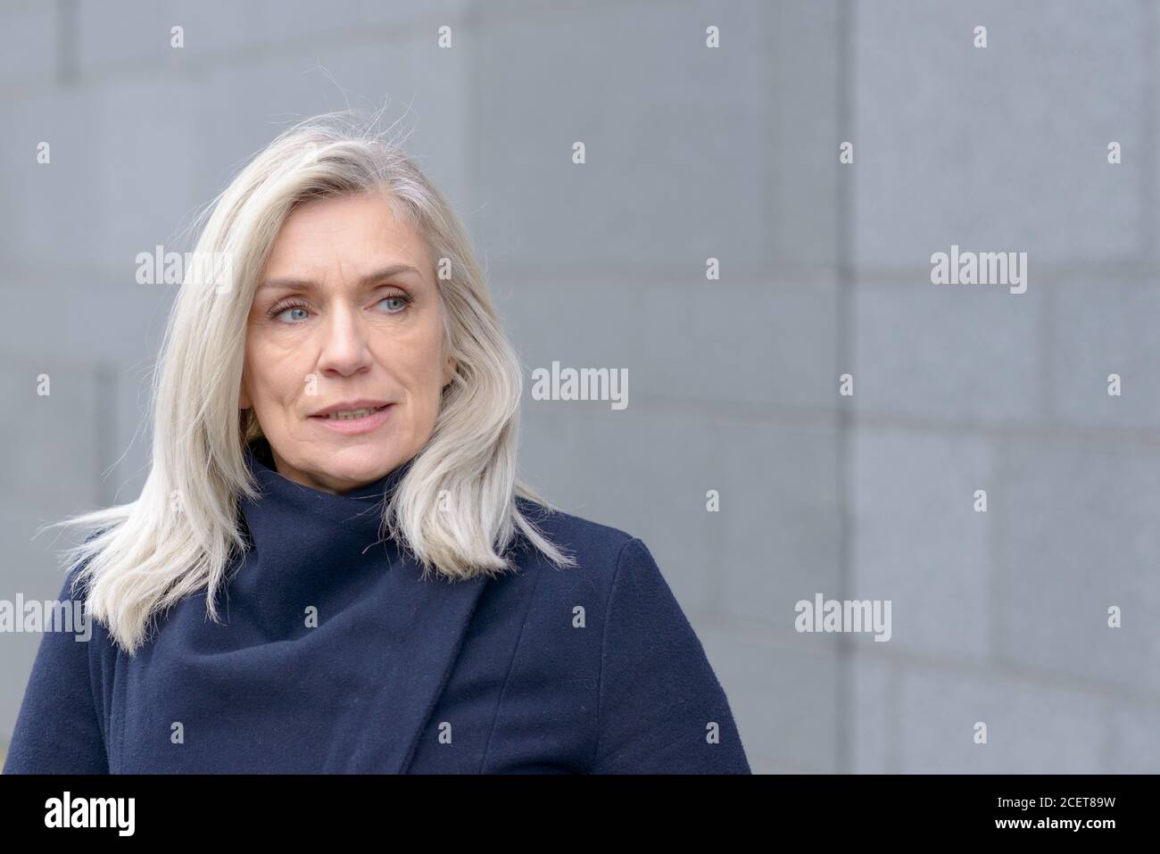 Nachdenklich attraktive blonde Frau beiseite, als sie draußen in einem warmen blauen Wintermantel gegen eine graue Wand Hintergrund steht Stockfoto
