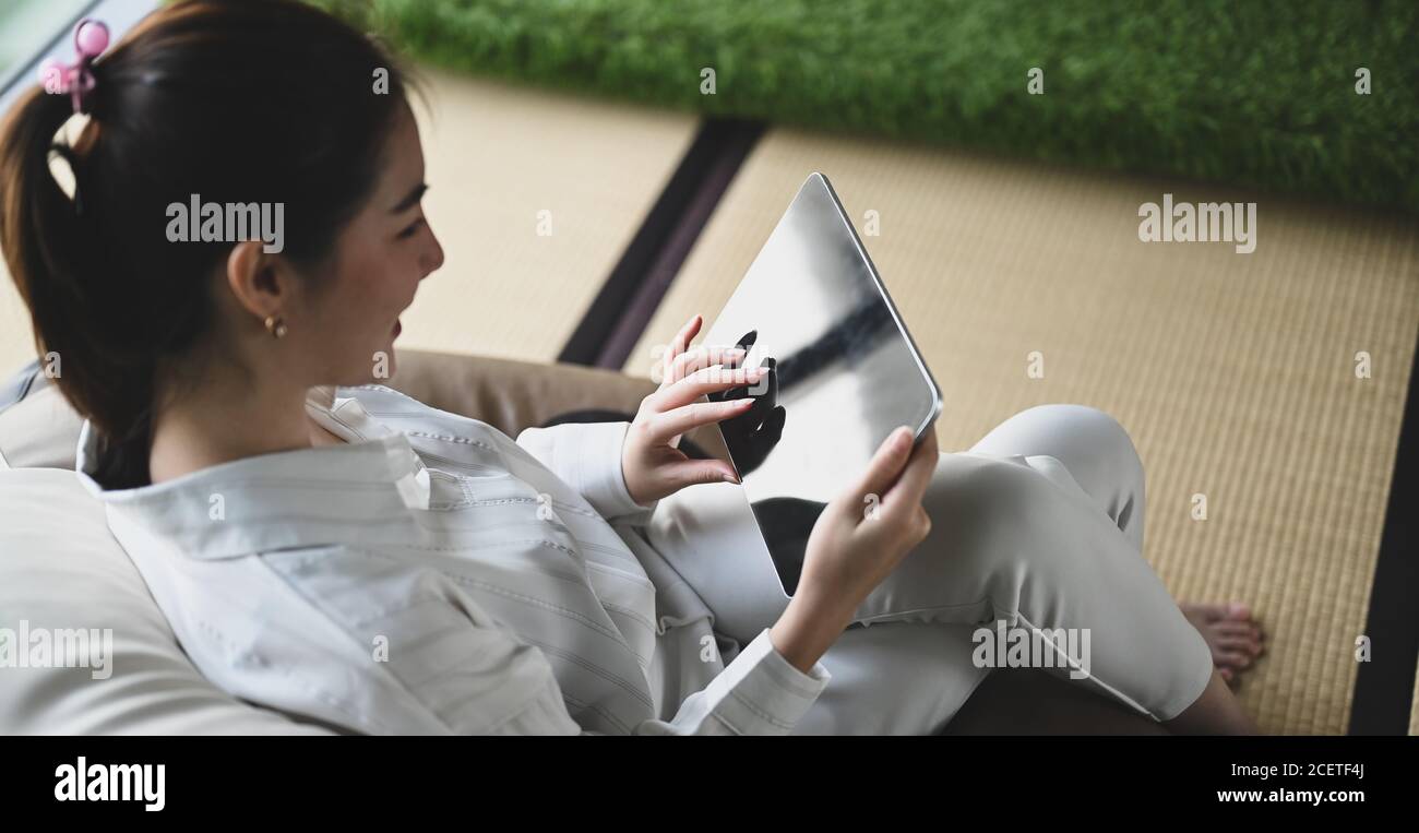 Eine junge Frau benutzt ein Computer-Tablet, während sie auf einem großen gepolsterten rahmenlosen Stuhl sitzt. Stockfoto