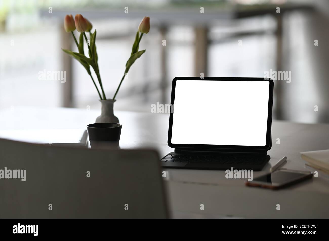 Ein Computertablett mit einem weißen leeren Bildschirm wird auf einen Schreibtisch gestellt, der von verschiedenen Geräten umgeben ist. Stockfoto