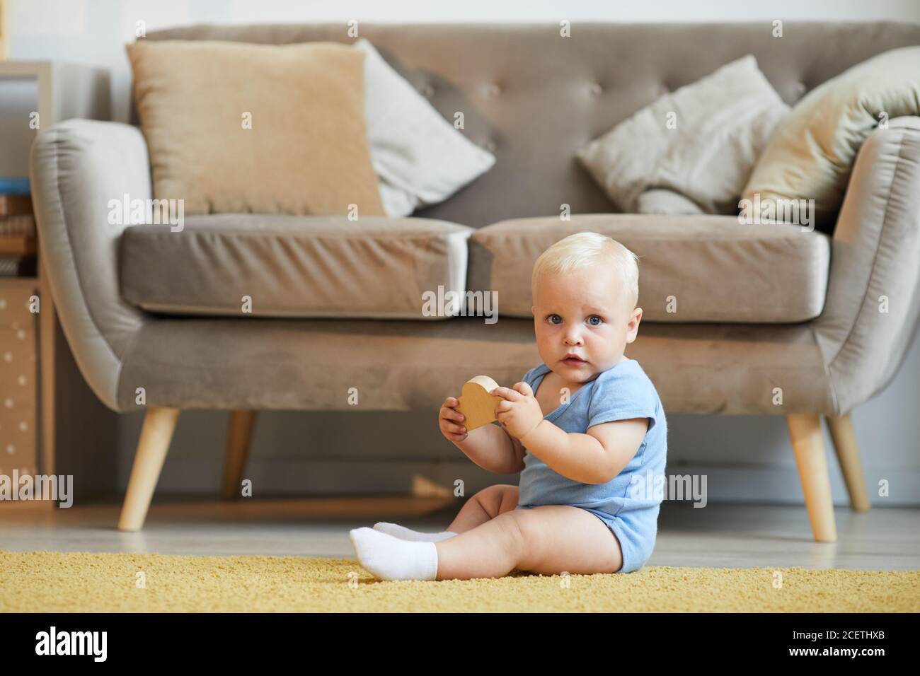 Horizontale lange Aufnahme des verspielten Babys, das auf dem Boden sitzt Wohnzimmer spielen mit Spielzeug Blick auf Kamera Stockfoto