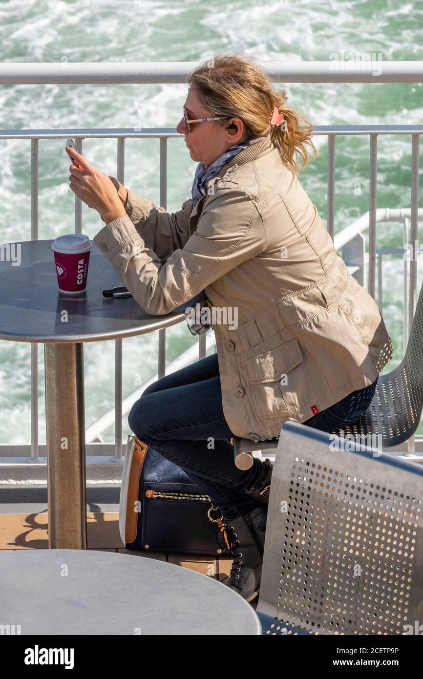 Eine junge attraktive Frau, die an einem Tisch auf einer Fähre oder einem Boot sitzt und ein mobiles Gerät oder Telefon anschaut und Kaffee aus einer Pappbecher trinkt. Stockfoto
