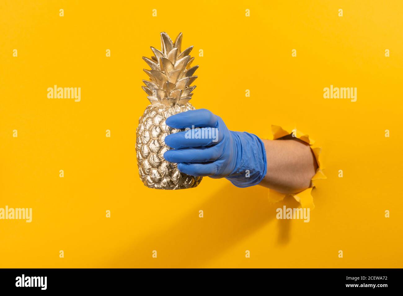 Hand in einen Latexhandschuh halten eine goldene Ananas durch Abgerissene gelbe Wand Stockfoto