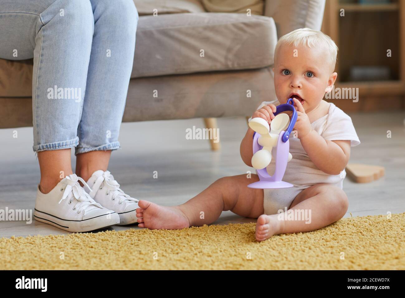 Horizontale Aufnahme des verspielten Babys, das auf dem Boden im Wohnzimmer sitzt und mit Spielzeug spielt, das die Kamera anschaut, seine Mutter auf dem Sofa sitzt Stockfoto