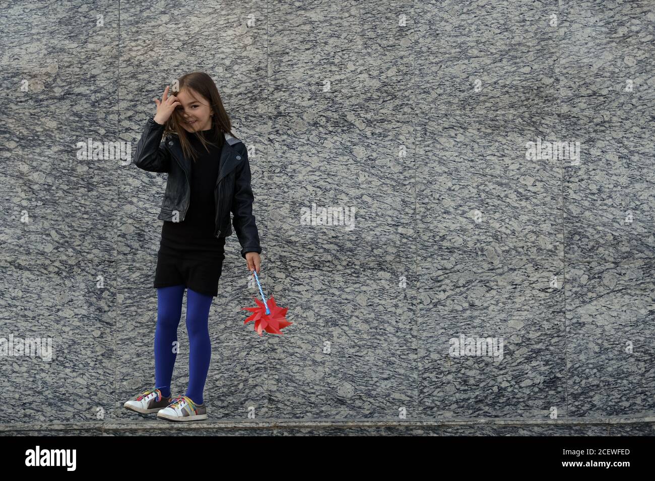 Kleine Stadt stilvolle Mädchen in lässigen Herbst Kleidung zu Fuß entlang graue Granitwand mit rotem Wind Spinner. Leerzeichen für Text. Stockfoto