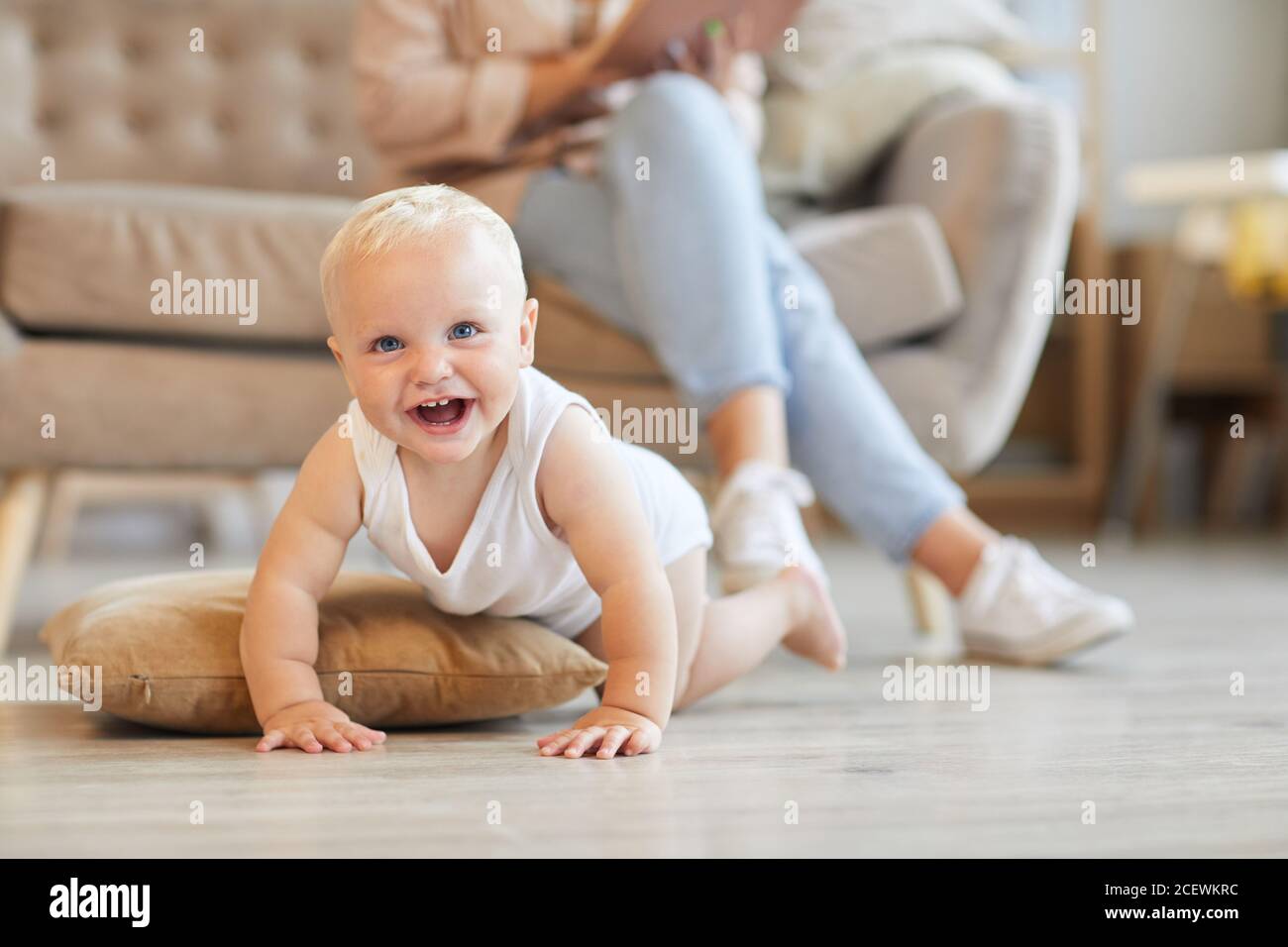 Horizontale Aufnahme von verspielten Baby Spaß auf dem Boden in Wohnzimmer, während seine Mutter auf dem Sofa sitzt und liest Buchen Stockfoto