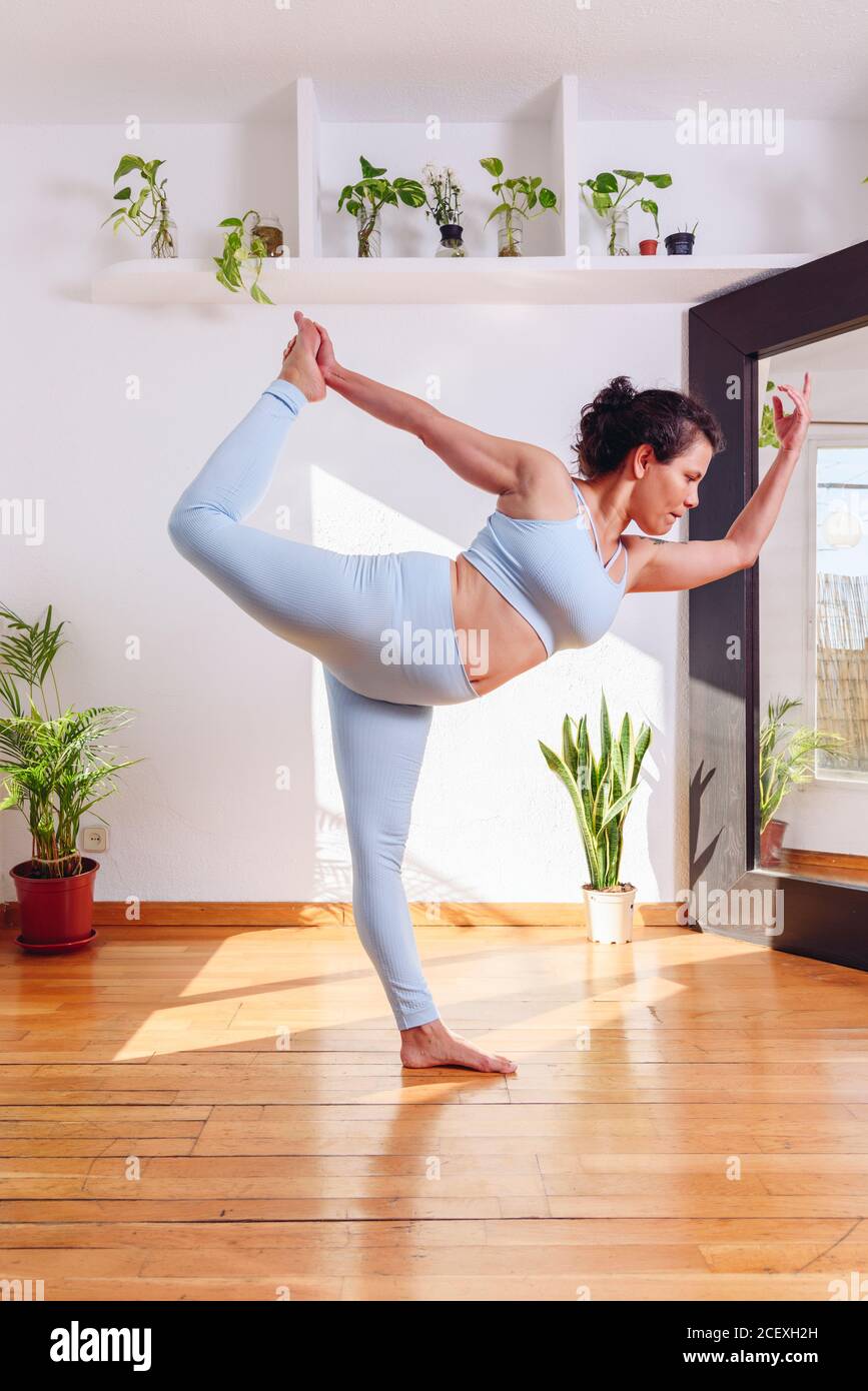Seitenansicht der fokussierten Frau in Sportbekleidung, die Yoga praktiziert Herr des Tanzes Pose, während sie auf Holzboden stehen Zu Hause Stockfoto