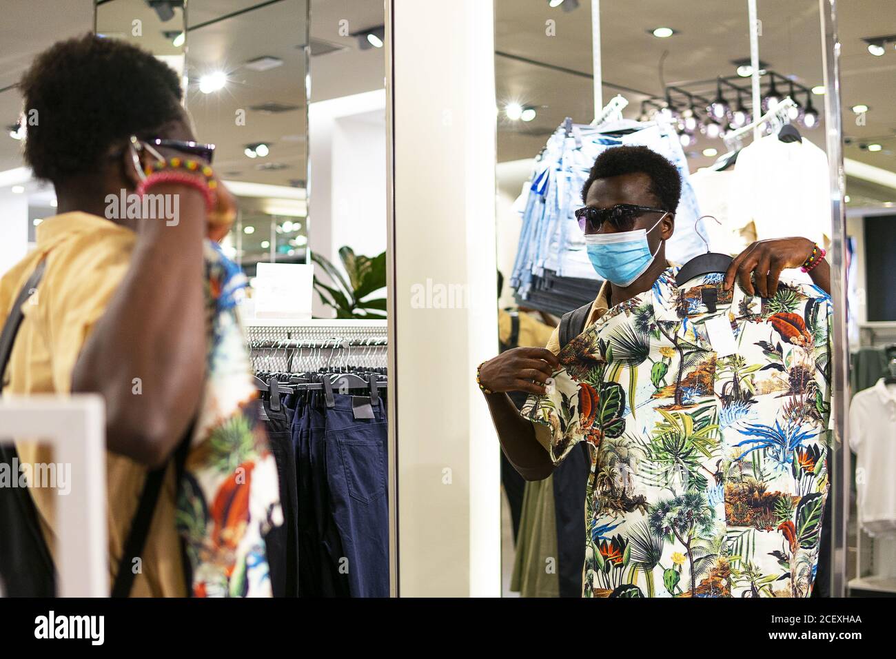 Junger afroamerikanischer Mann in trendigem Outfit und medizinischer Maske Vor dem Spiegel in der modernen Boutique stehen und pflücken Ware im Laden während Coronavirus Stockfoto
