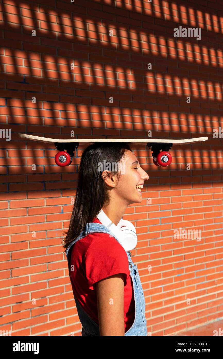 Seitenansicht der weiblichen Millennial stehend mit Skateboard auf dem Kopf In der Nähe der Ziegelmauer und lachen, während Spaß haben Stockfoto