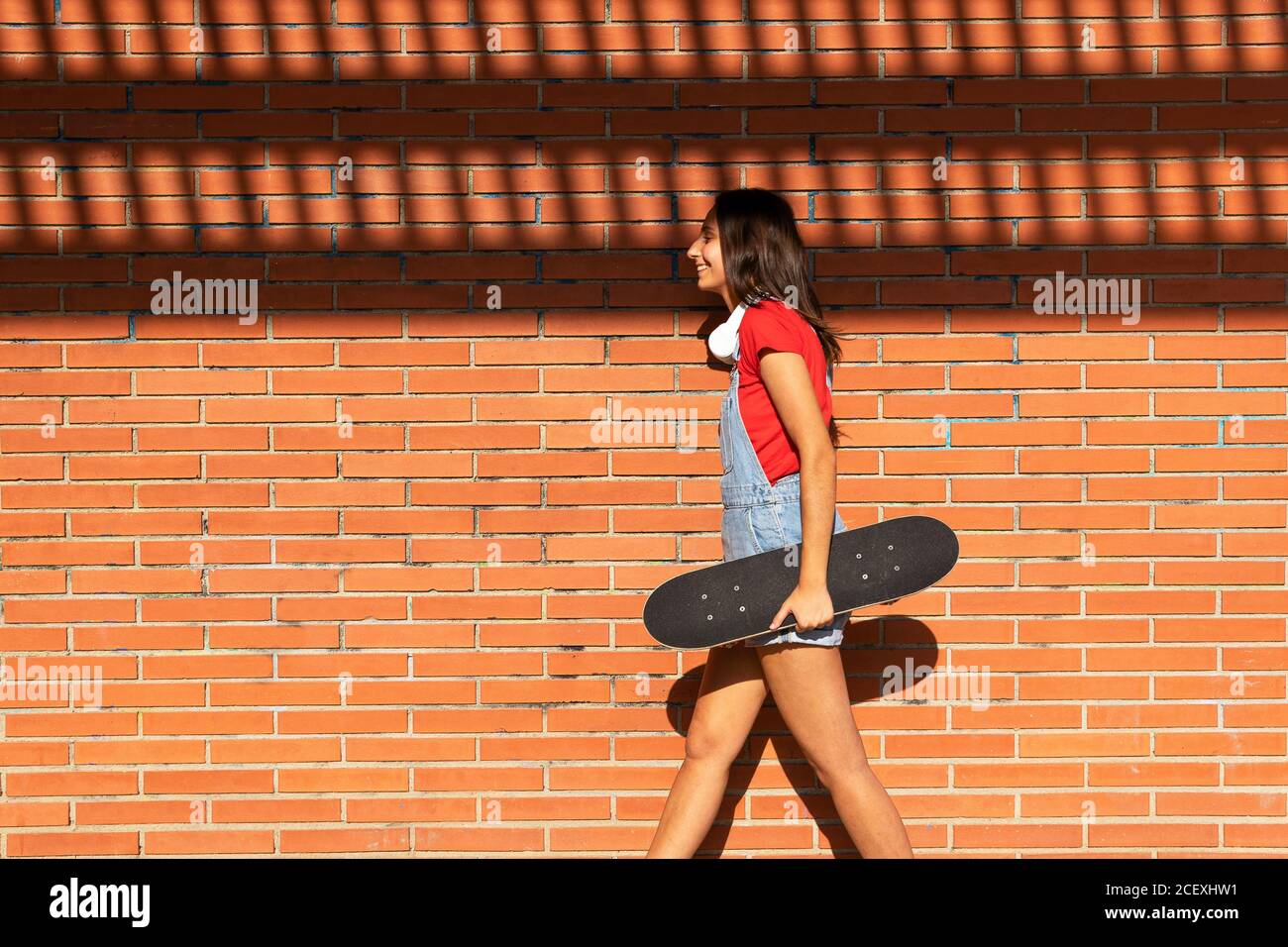 Seitenansicht des weiblichen Millennial Walking mit Skateboard in der Nähe von Backstein Wand und lachen, während Spaß haben Stockfoto