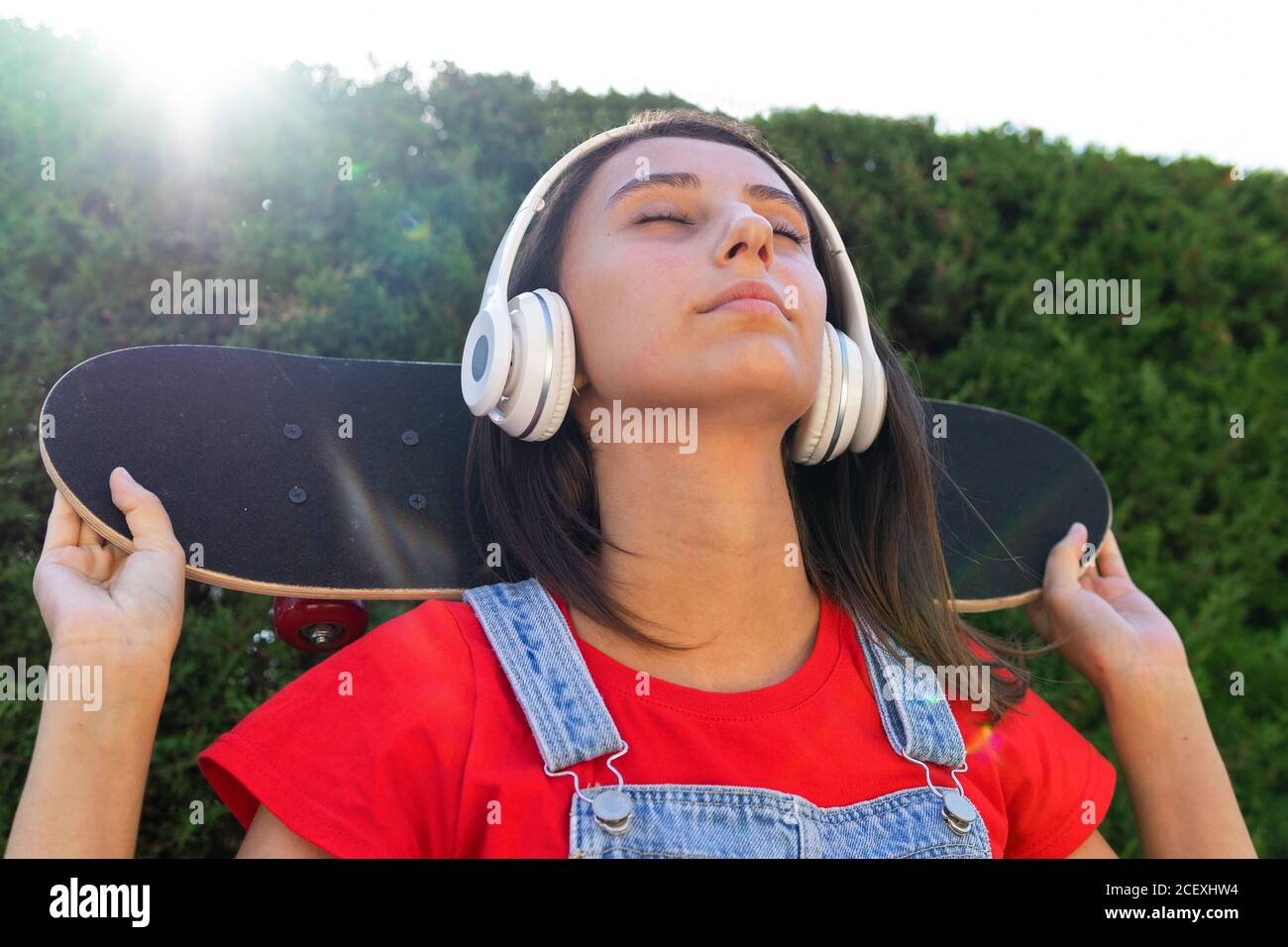 Seitenansicht der weiblichen Millennial stehend mit Skateboard auf dem Kopf In der Nähe der Ziegelmauer und lachen, während Spaß haben Stockfoto