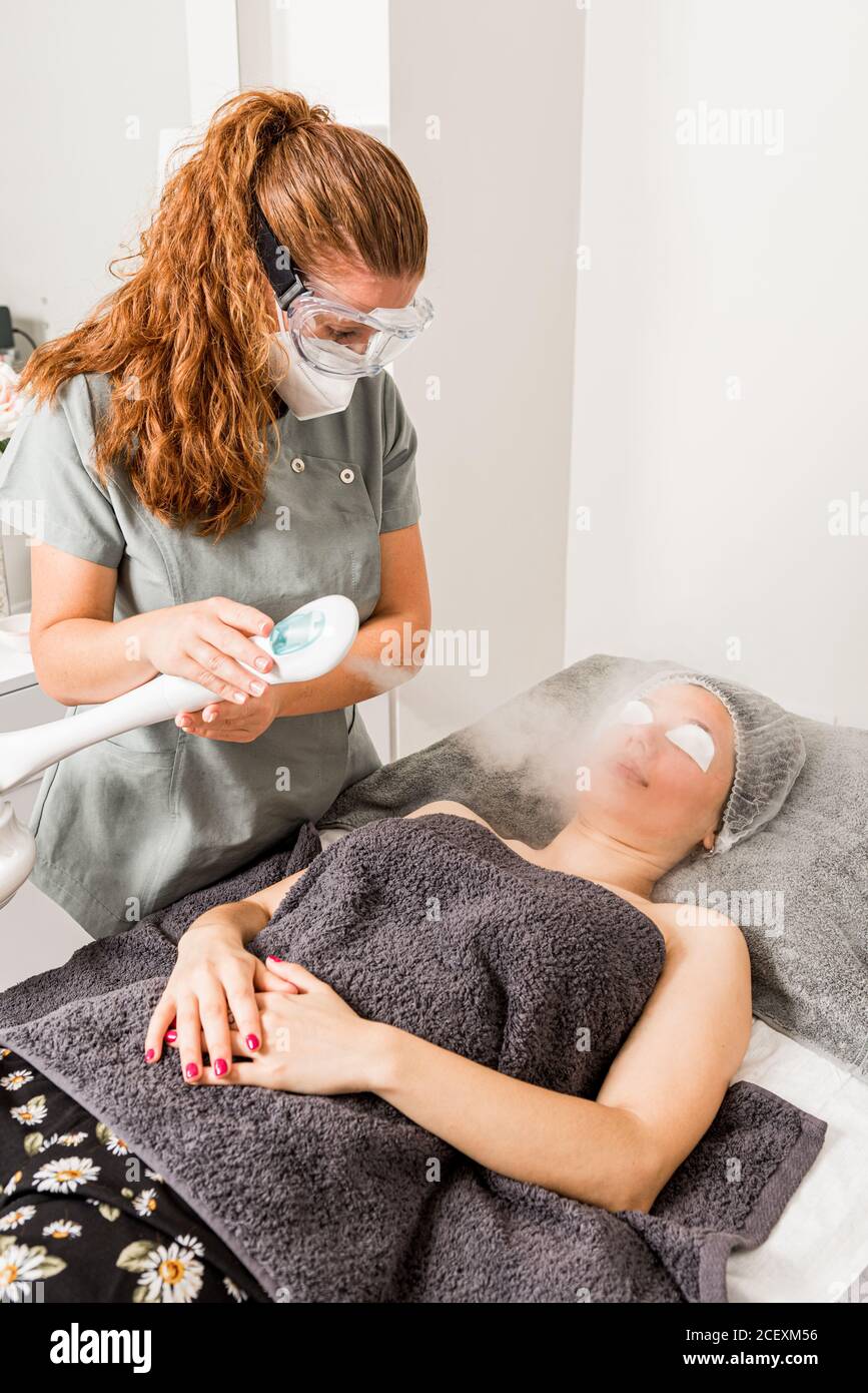 Professionelle Schönheitstherapeut in Schutzmaske und Schutzbrille mit Gesichtsbehandlung Dampfgarer für die Reinigung Behandlung von weiblichen Kunden in der zeitgenössischen Kosmetologie Klinik Stockfoto