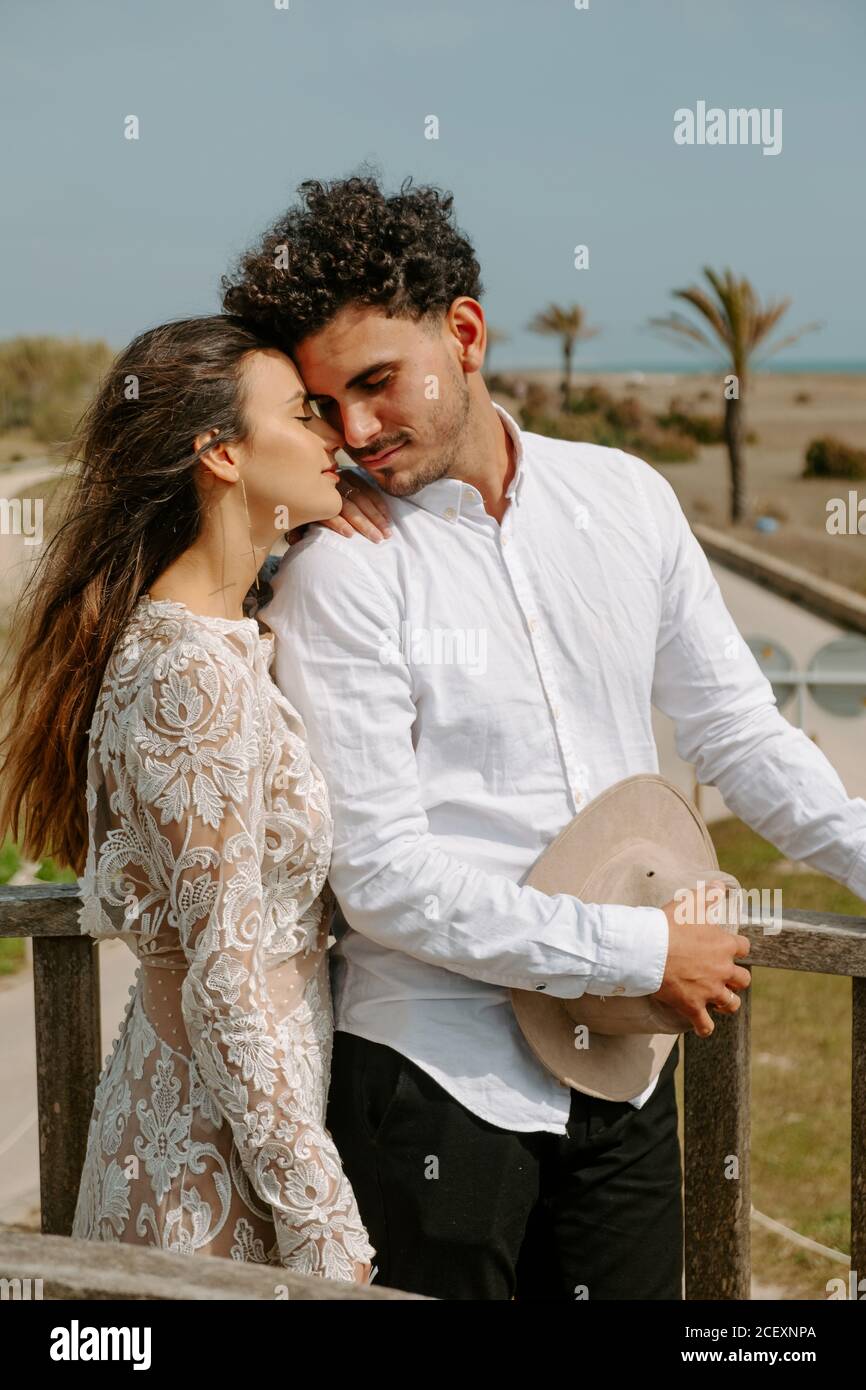 Seitenansicht eines entspannten jungen Paares in stilvollen weißen Outfits Stehen auf Holzbrücke mit geschlossenen Augen kuscheln einander Während Sie einen romantischen Urlaub am Meer verbringen Stockfoto