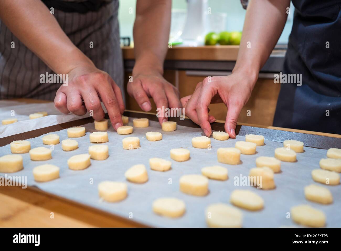 Zugeschnittene, nicht erkennbare Hände von Mann und Frau in Schürzen, die in der Bäckerei zu Hause arbeiten und Cookies auf Backform mit Pergament legen Stockfoto