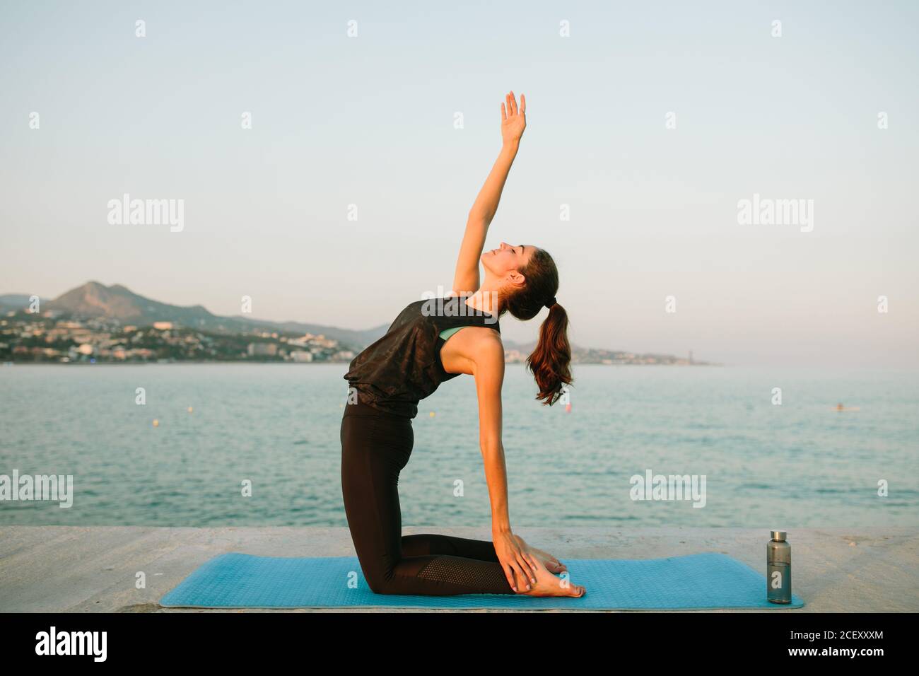 Seitenansicht einer ruhigen Frau in Ustrasana, die Yoga praktiziert Geschlossene Augen und erhobener Arm auf Pier in der Nähe des Meeres während Sonnenuntergang im Sommer Stockfoto