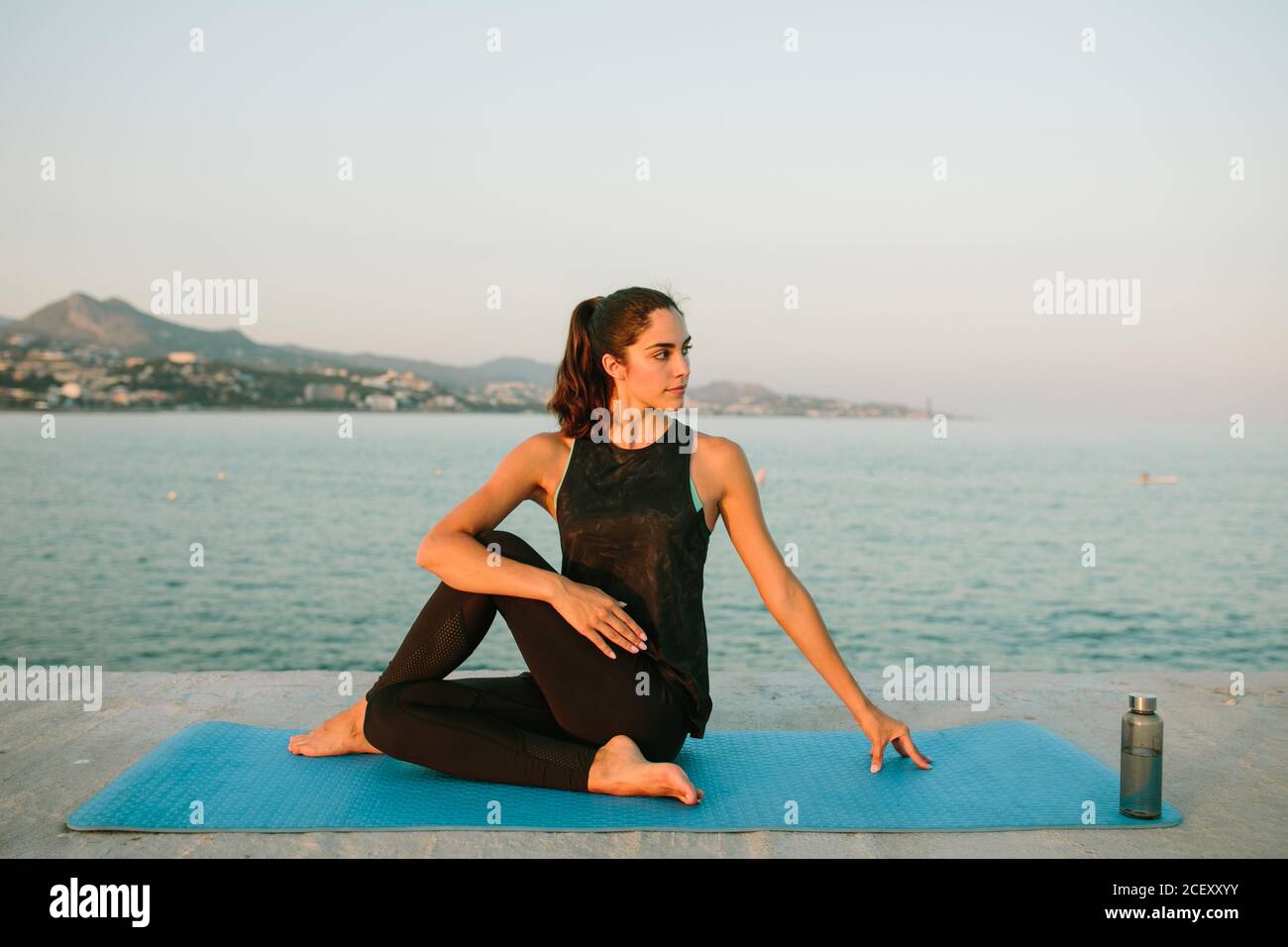 Junge unbeschwerte Frau sitzt auf Yoga-Matte in Ardha Matsyendrasana Und Stretching Körper beim Blick weg auf den Hintergrund der Seetang Abends Stockfoto