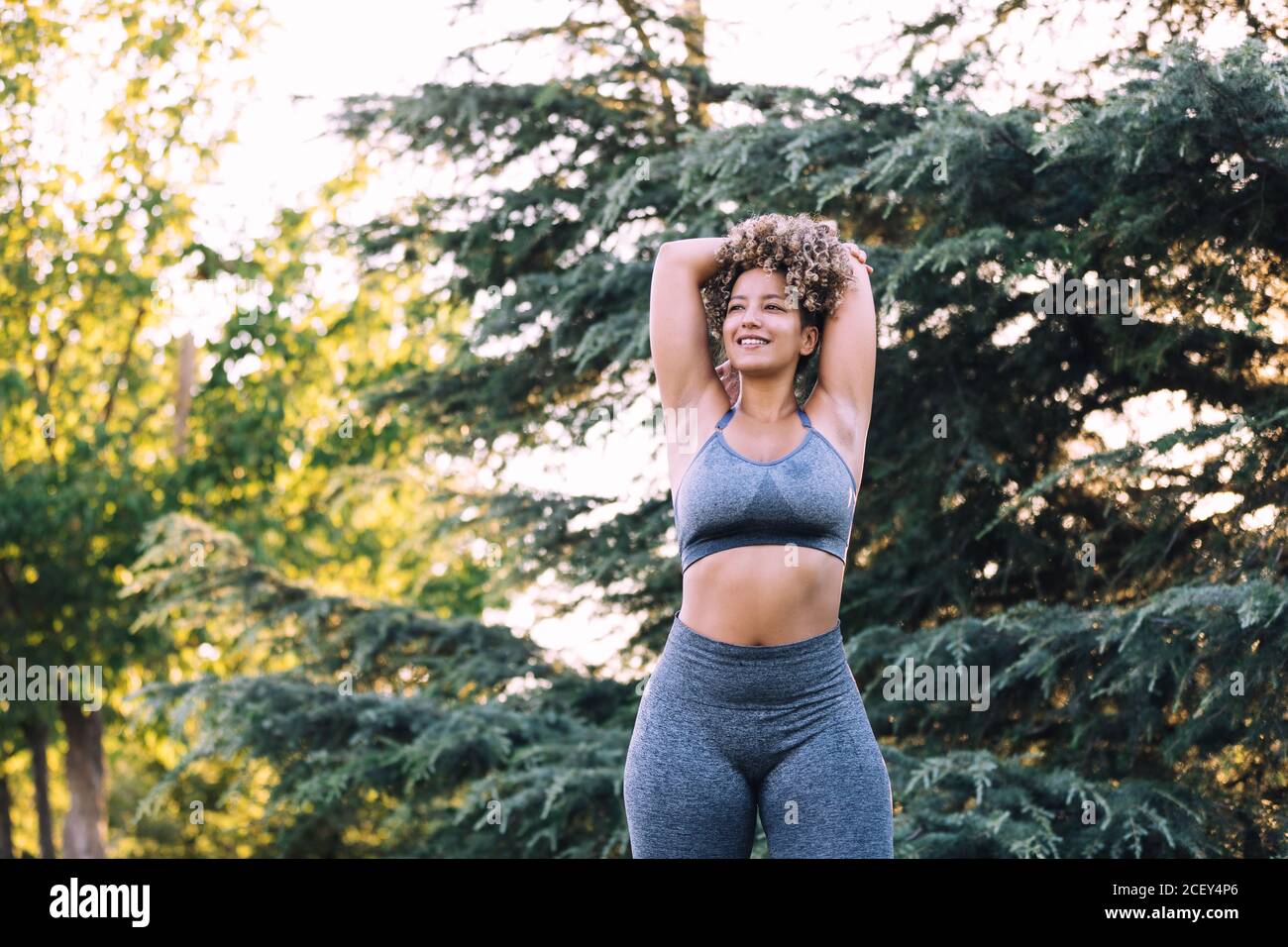 Niedriger Winkel der positiven energischen jungen ethnischen weiblich mit Afro Frisur mit grauem Oberteil und Leggings mit Armen hinten Kopf während des Trainings im Park in der Nähe von grünen Bäumen stehen Stockfoto