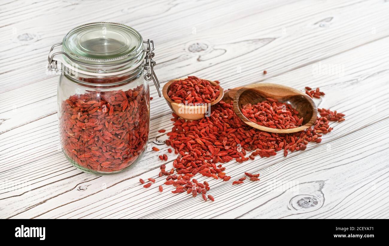 Getrocknete Goji aka. wolfberry Samen in Glasflasche, einige in Holzschüsseln und verschüttet auf weißen Brettern Schreibtisch Stockfoto