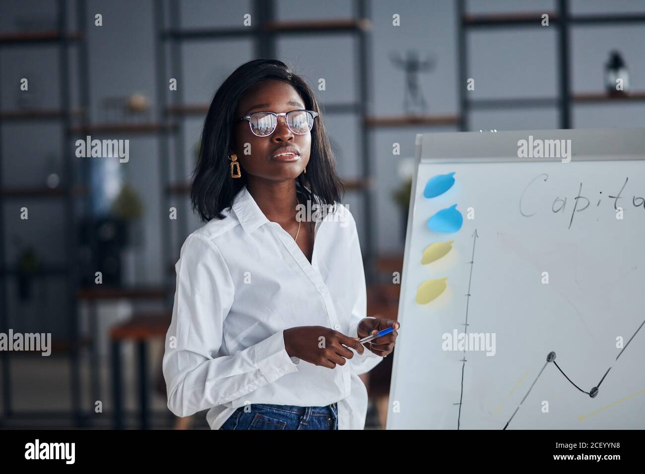 Selbstbewusste afrikanische Frau in Brillen, trägt weißes Hemd und Jeans, mit Diagrammen, motivierte afroamerikanische Trainer präsentieren Business-Plan o Stockfoto