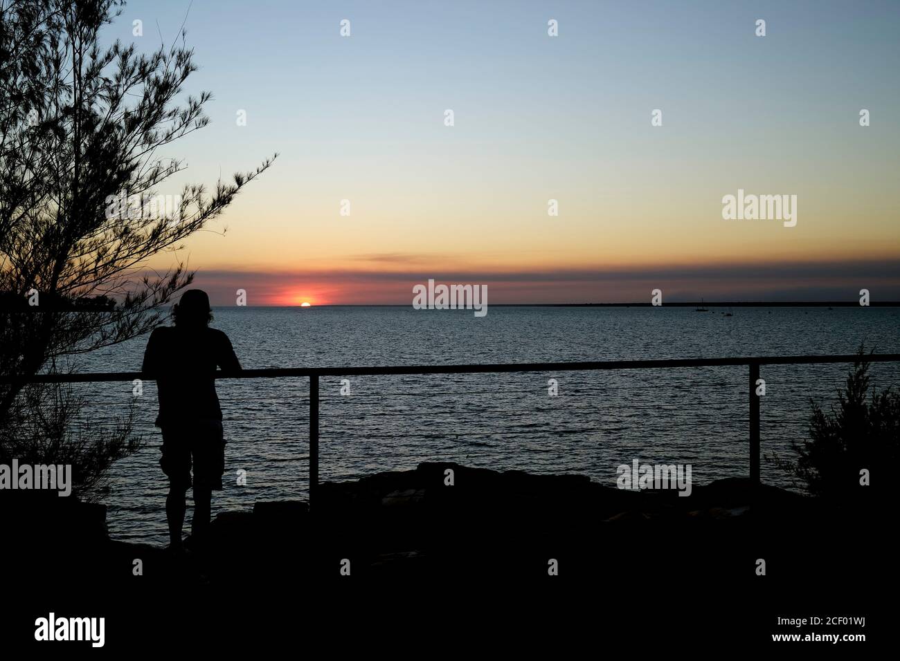 Silhouette eines Mannes, der sich auf einen Zaun stützt, um den Sonnenuntergang über einem großen Gewässer zu beobachten. Stockfoto
