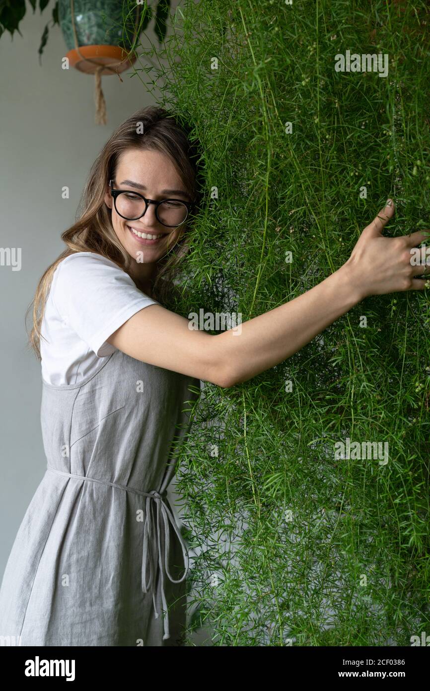 Glückliche junge Frau Gärtner in Pfandrecht Kleid, umarmt üppigen Spargel Farn Zimmerpflanze in ihrem Blumengeschäft. Viel Grün zu Hause. Liebe zu Pflanzen. Innen gemütlich Stockfoto
