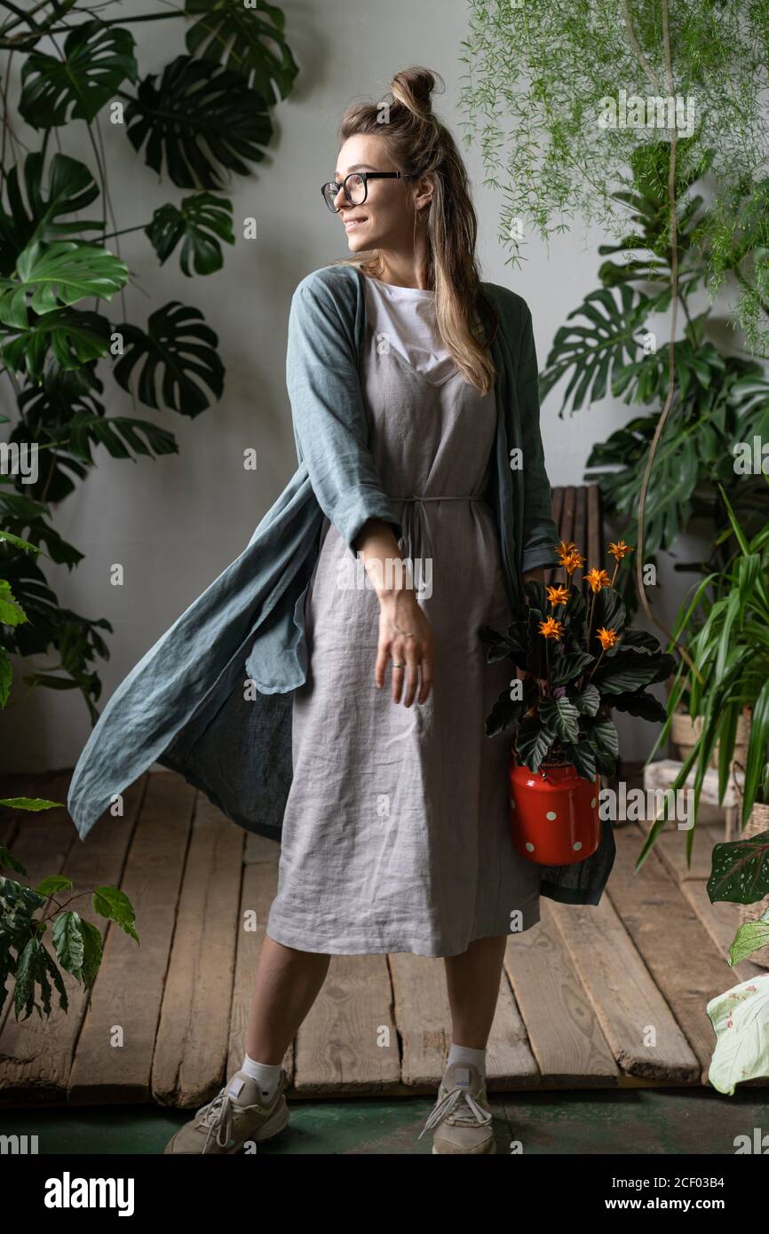 Glückliche Frau Gärtnerin in Brillen tragen Leinenkleid, hält eine blühende calathea Pflanze in alten roten Milch kann Blick auf Fenster, stehend in ihrem Haus Stockfoto