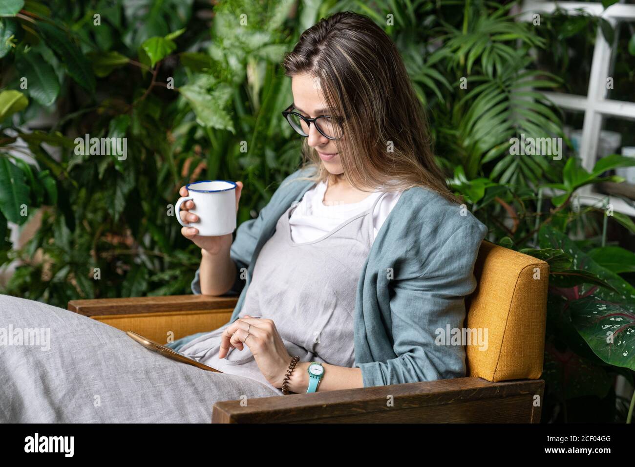 Die Gärtnerin trägt Leinenkleid, sitzt auf einem Stuhl im grünen Haus, ruht sich aus, benutzt Smartphone und trinkt Tee aus einer Tasse in Harmonie mit den Pflanzen ar Stockfoto