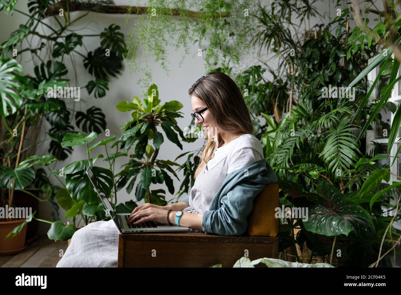 Nahaufnahme Porträt einer lächelnden Gärtnerin auf einem Stuhl im Gewächshaus sitzen, mit Laptop und sprechen im Web-Anruf von exotischen Pflanzen umgeben. Stockfoto