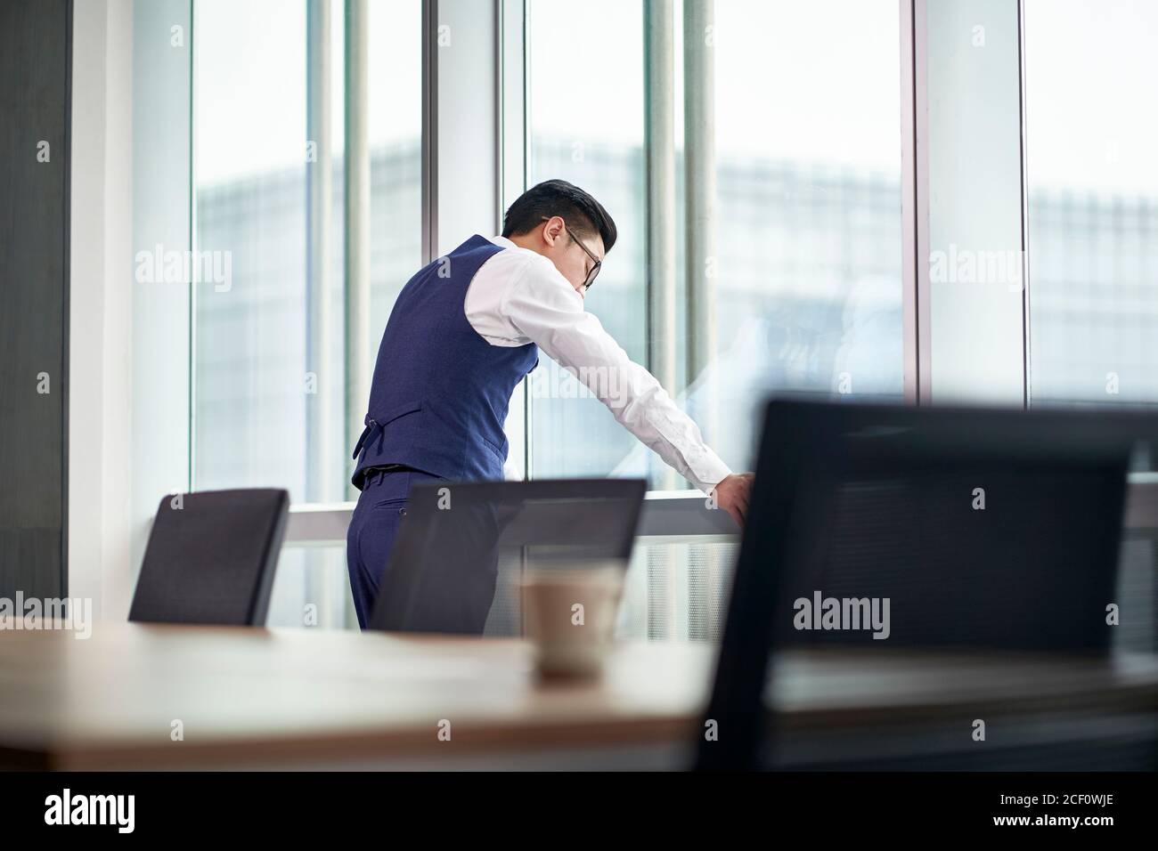 Frustrierter asiatischer Geschäftsmann, der im Büro am Fenster stand, den Kopf nach unten und die Augen geschlossen Stockfoto