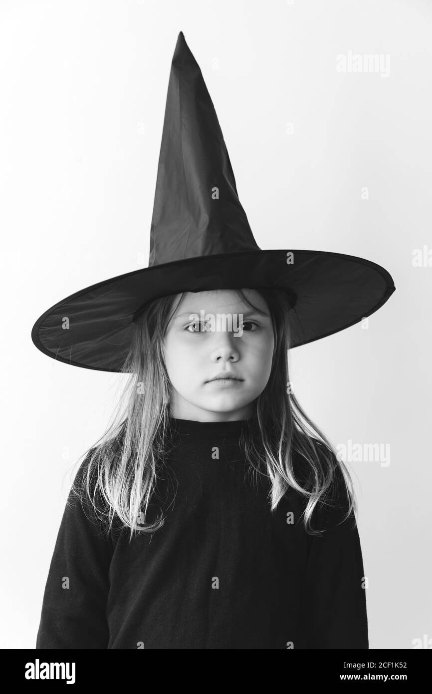 Kleines Mädchen im schwarzen Kostüm mit Hexenhut steht über weißer Wand, schwarz-weiß stilisiertes Foto Stockfoto