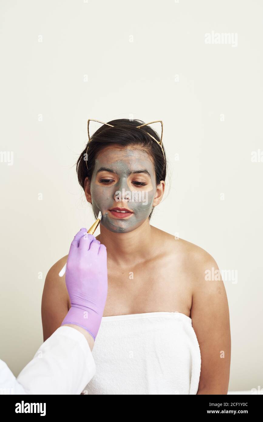Eine graue Schlammmaske wird auf eine schöne junge Frau aufgetragen. Schönheitsbehandlungen. Gesichtspflege, Poren straffen und feuchtigkeitsspendend mit natürlichen kosmetischen Cremes. Stockfoto