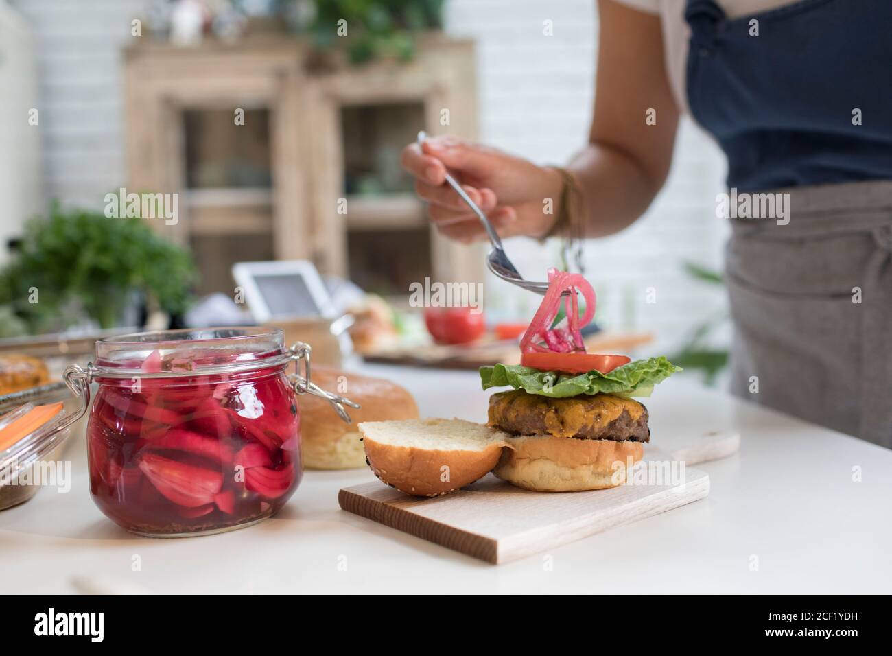 Frau, die in der Küche eingelegte Zwiebeln über Cheeseburger legt Stockfoto