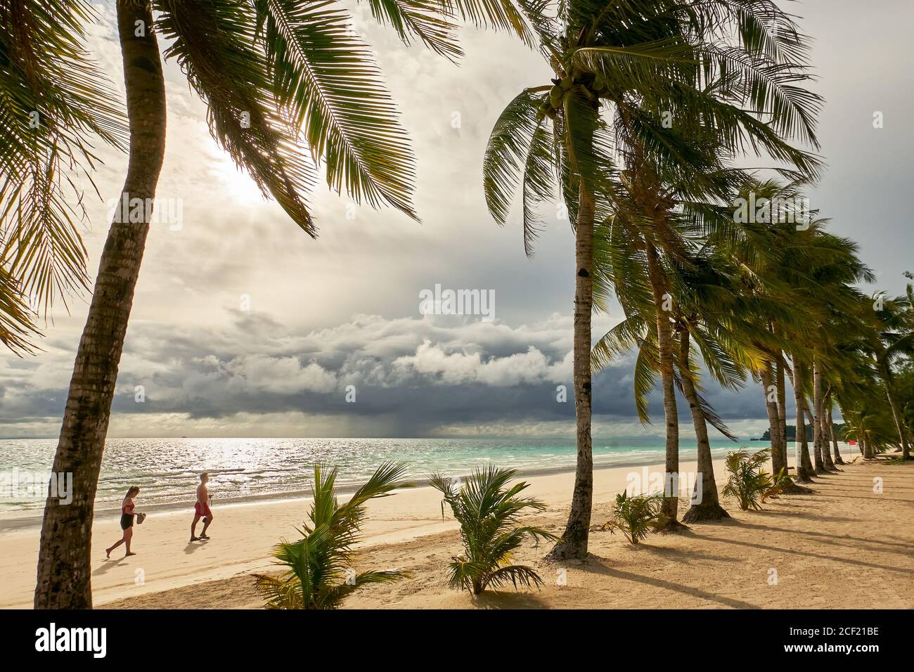 Junges Paar, das am sauberen weißen Strand mit Kokospalmen bei Sonnenuntergang mit bewölktem Himmel auf Boracay Island, Provinz Aklan, Visayas, Philippinen, spazierengeht Stockfoto