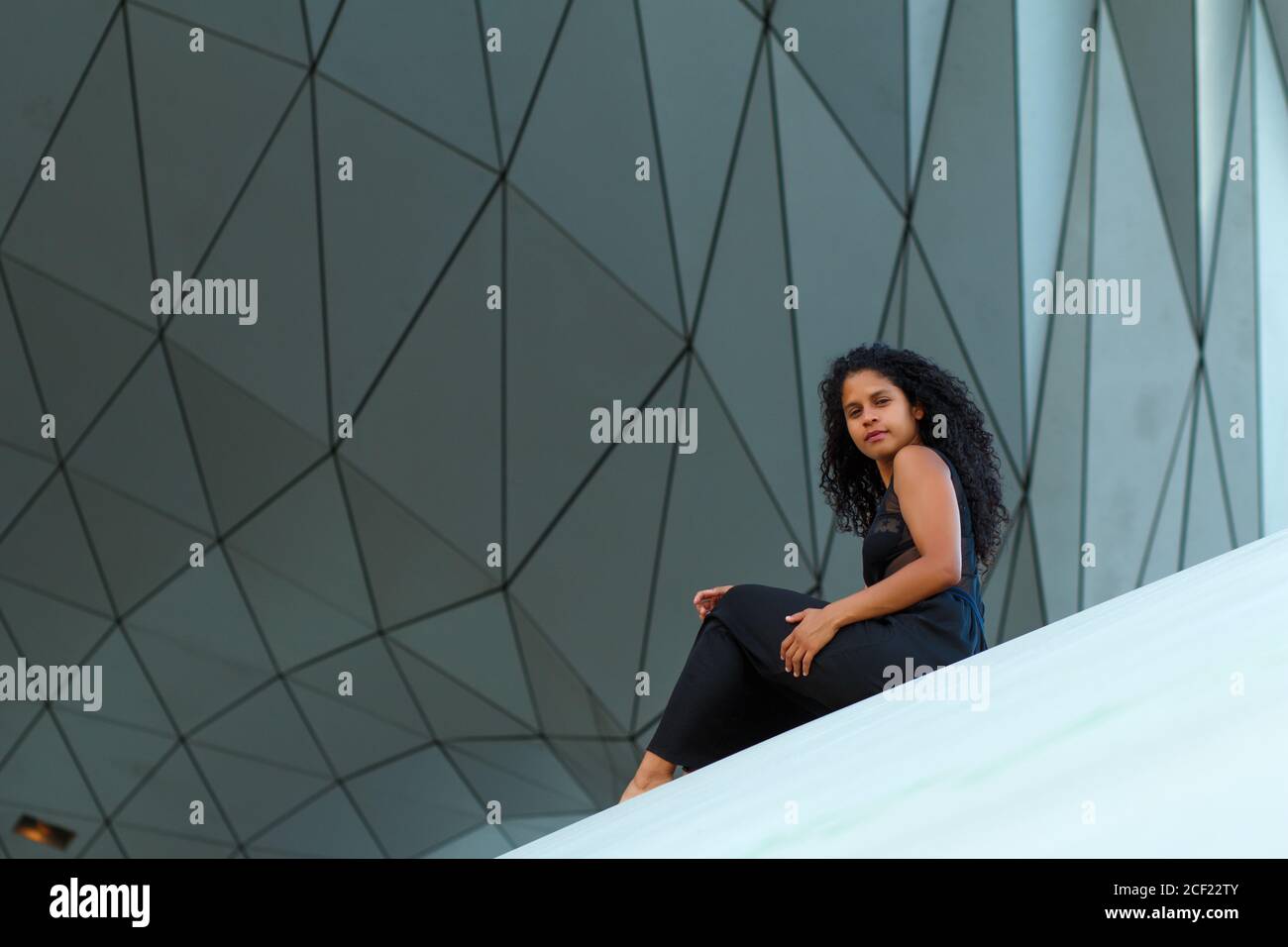 Lockige Brünette Hispanic weiblich ein elegantes schwarzes Kleid sitzt unter Die Wand eines modernen Gebäudes Stockfoto