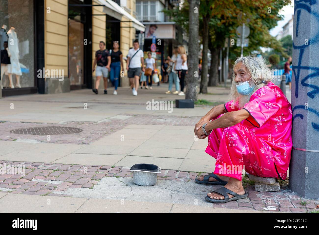 Alter extravaganter bärtiger Mann mit langen weißen Haaren und Gesicht Maske trägt saubere rosa Kleidung betteln für Geld in der Straßen von Sofia Bulgarien Stockfoto