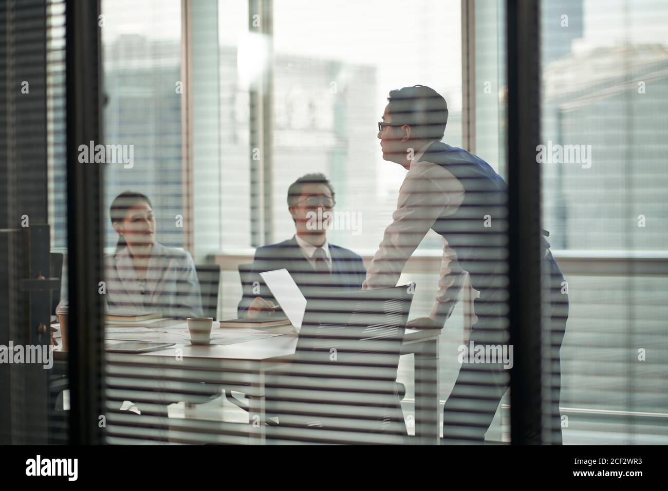 Durch-das-Glas-Aufnahme eines asiatischen Managers, der oben steht Der Teammitglieder, die am Besprechungstisch sprechen Stockfoto