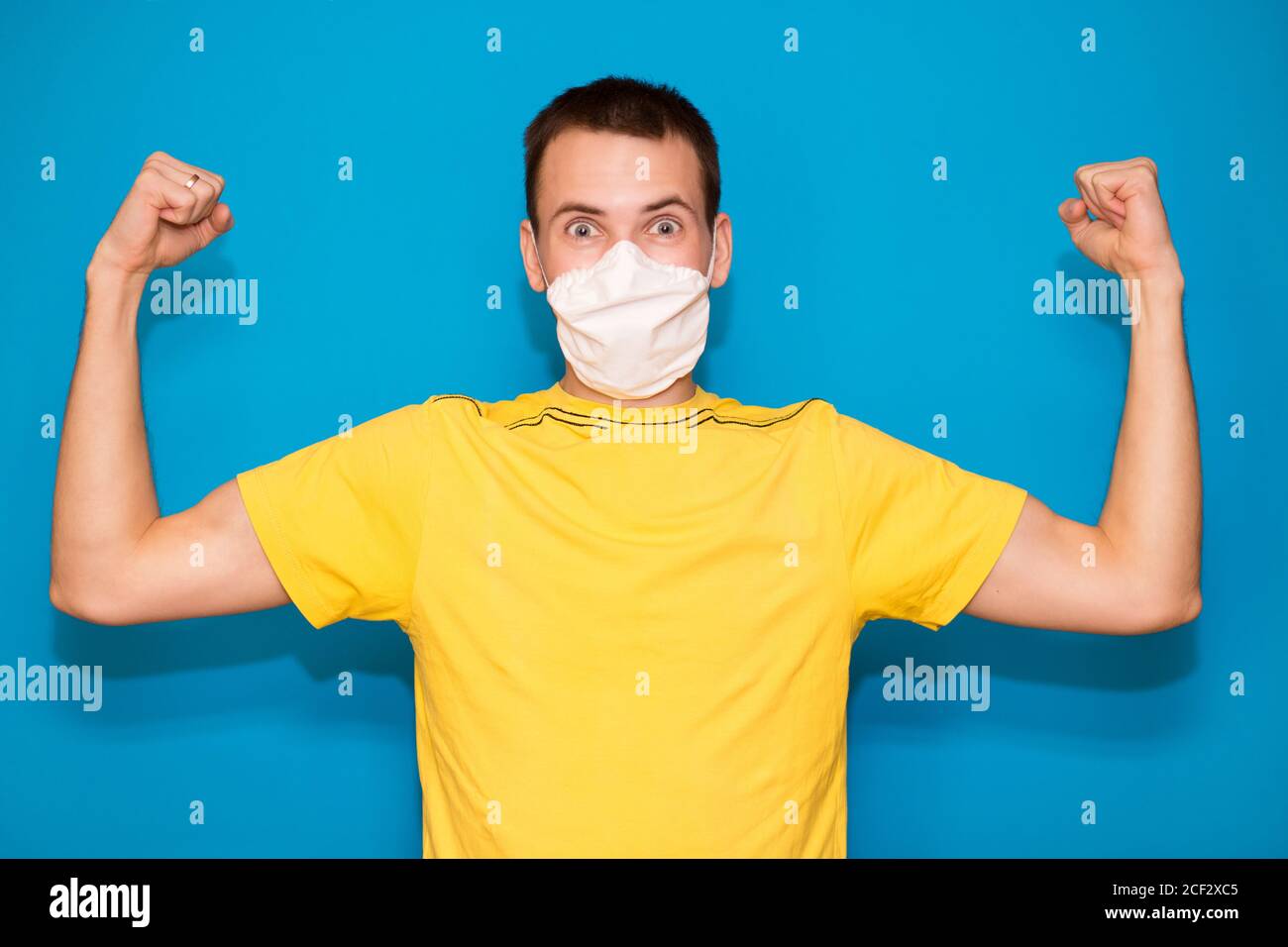 Junger gutaussehender Mann in medizinischer Maske und gelbem T-Shirt isoliert auf blauem Hintergrund. Starke Person zeigt Arm Muskel, zuversichtlich und stolz auf die Macht zu d Stockfoto