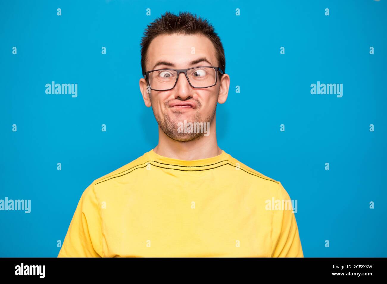 Lustige Comic-Mann in Brille macht Grimasse mit Kreuzen Augen. Junger Mann in gelbem T-Shirt mit verrücktem Ausdruck hat Spaß allein, spielt Narr isoliert über Stockfoto