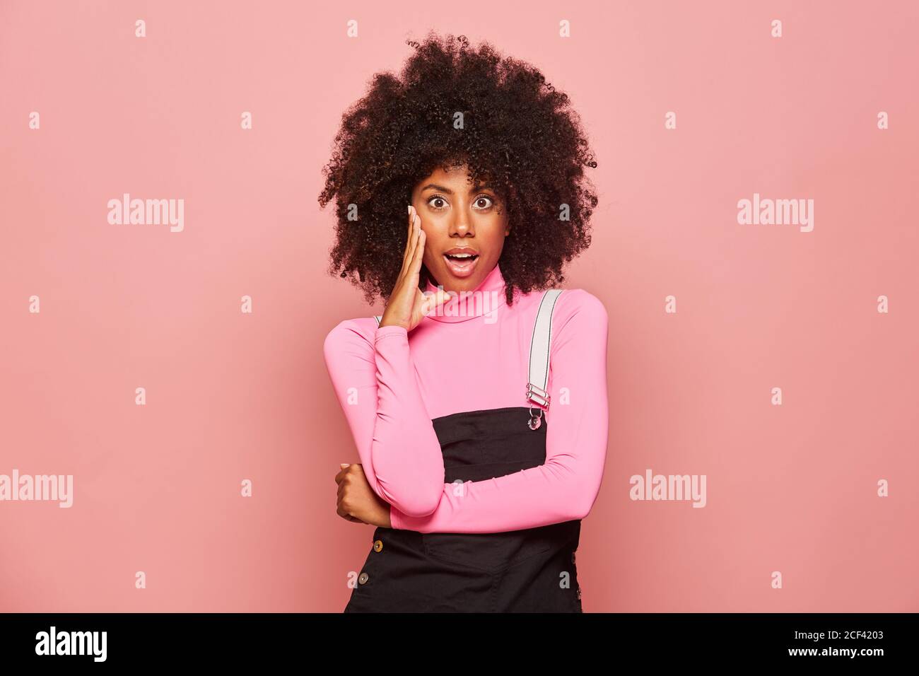 Erstaunt afroamerikanische Frau trägt rosa Rollkragen und schwarz insgesamt Das Gesicht berühren und die Kamera mit offenem Mund betrachten überrascht Mit Nachrichten Stockfoto