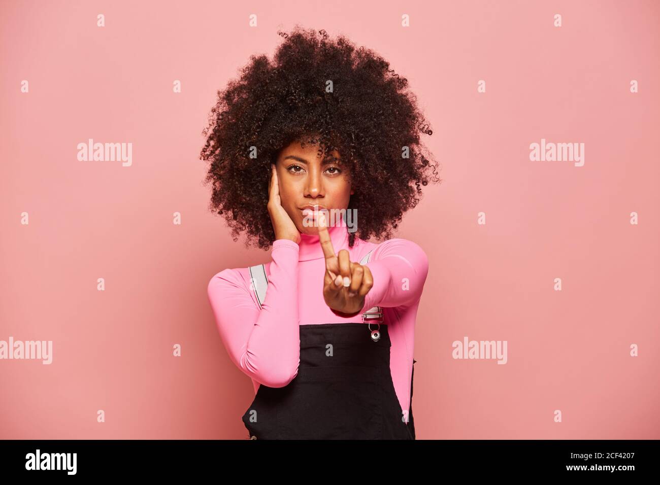 Junge afroamerikanische Frau mit lockigem Haar in rosa Rollkragen Und schwarz Overall hält Hand auf Wange und zeigt Nein Geste mit dem Finger nach oben, während er vor rosa Hintergrund steht Stockfoto