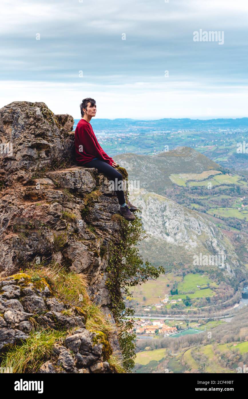 Seitenansicht der Touristen, die am Rand der Klippe sitzen und genießen Freiheit und bewundern erstaunliche Landschaft der Landschaft im Tal Am Bergvorhang gegen neblige bewaldete Hügel und Ebene unter Himmel mit üppigen grauen Wolken in Spanien Stockfoto