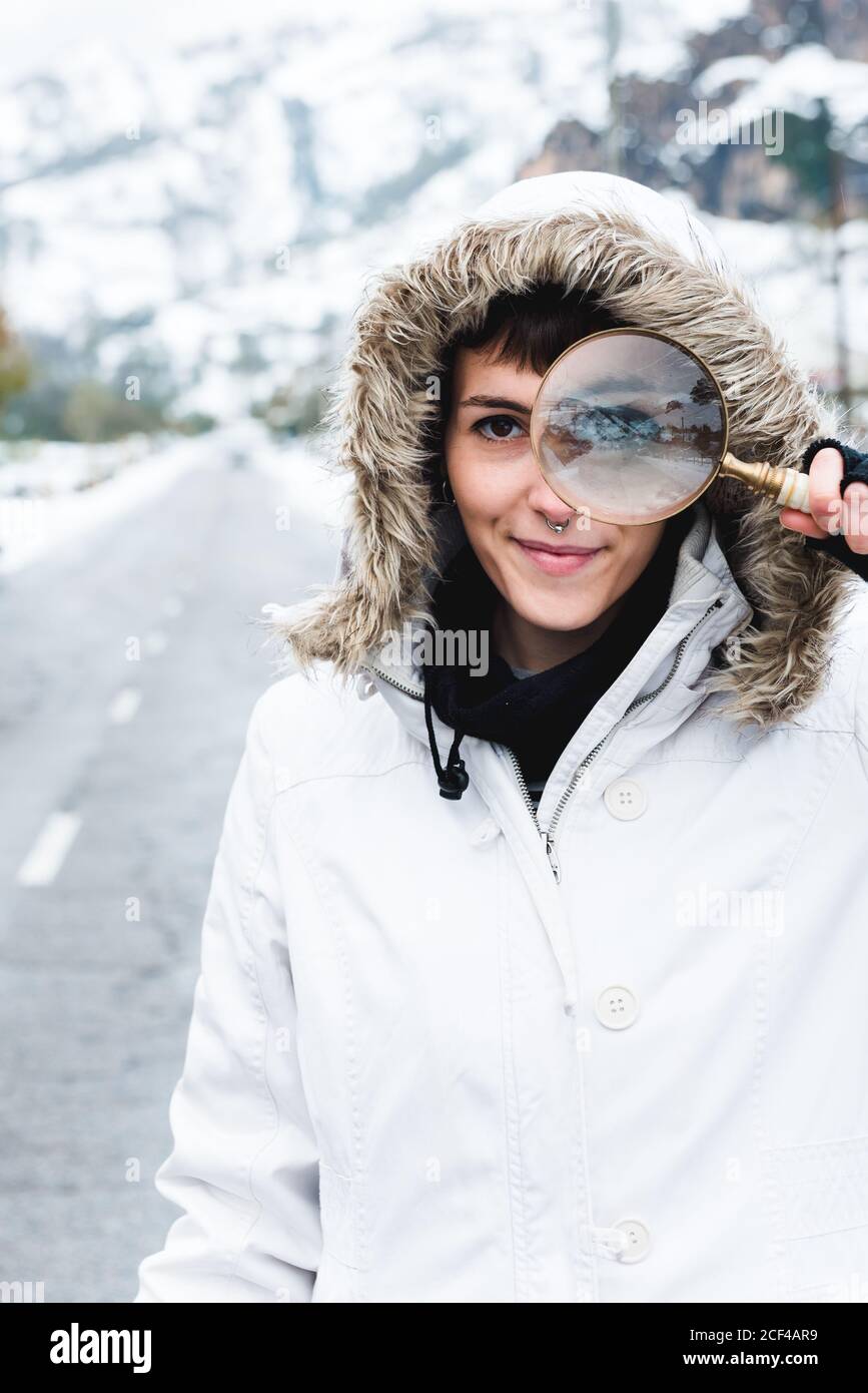 Inhalt interessierte Frau mit Nasenpiercing trägt weiße Winterjacke Mit Kapuze auf dem Kopf Blick auf die Kamera durch Lupe Während Sie auf einer leeren Straße in der Nähe von verschneiten Bergen stehen Stockfoto