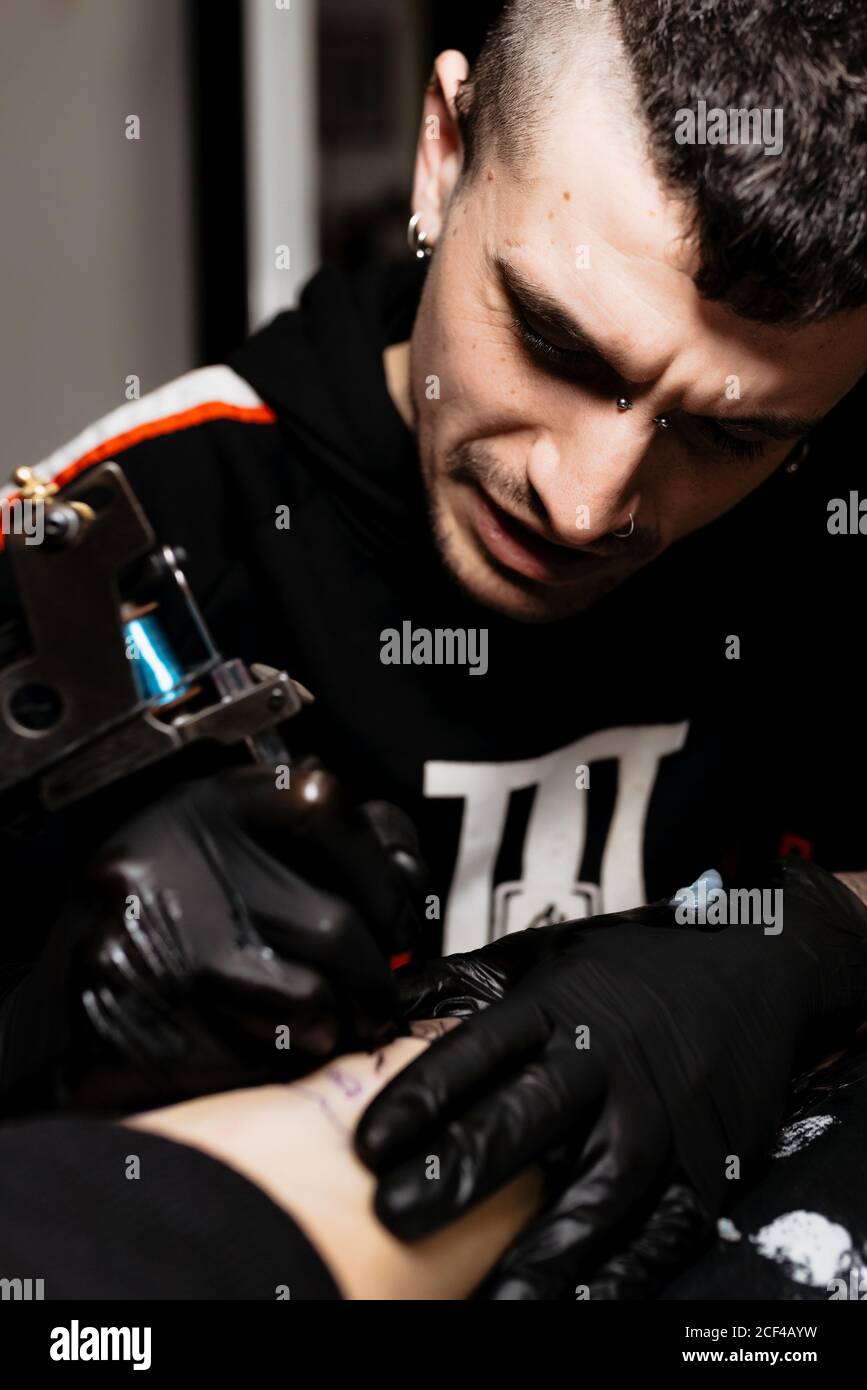 Stilvolle Mann mit Piercing mit Tattoo Maschine Tattoo zu machen Am Bein des Ernteguts Kunde während der Arbeit im Salon Stockfoto