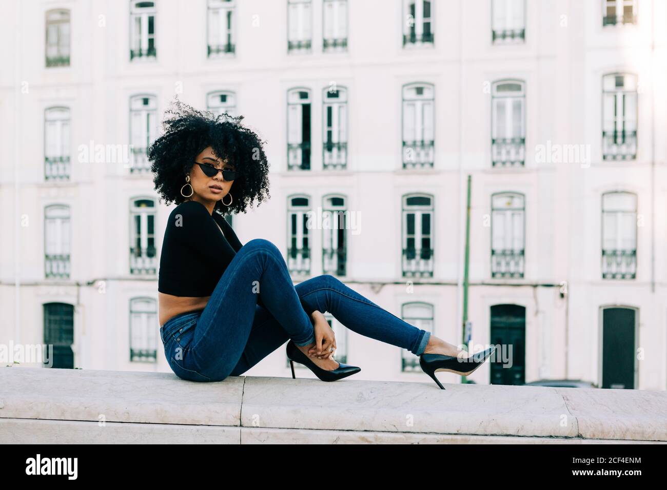 Junge trendige afroamerikanische Frau in Jeans und Crop Top sitzt auf Steinbrüstchen und schaut über Sonnenbrillen Stockfoto