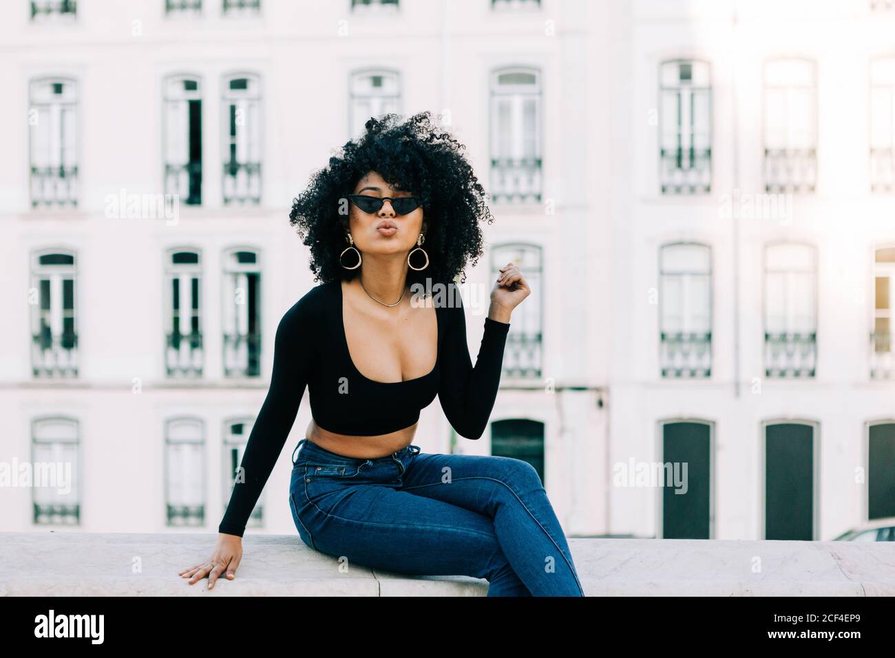 Junge trendige afroamerikanische Frau in Jeans und Crop Top sitzt auf Steinbrüstchen und schaut über Sonnenbrillen Stockfoto