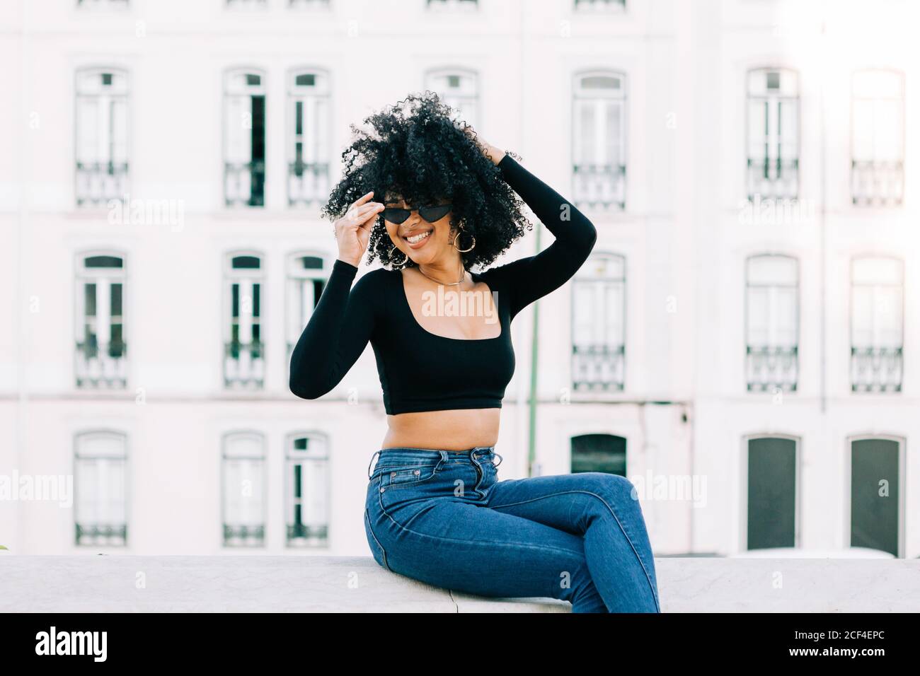Junge trendige afroamerikanische Frau in Jeans und Crop Top sitzt auf Steinbrüstchen und schaut über Sonnenbrillen Stockfoto