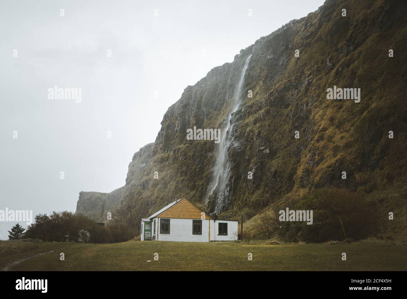 Kleines Landhaus aus Holz mit weißen Wänden und gelbem Giebeldach Dach auf grüner Wiese am Fuß der Klippe mit Wasserfall gegen grau bewölkten Himmel im Frühling Tag im Norden Irland Stockfoto
