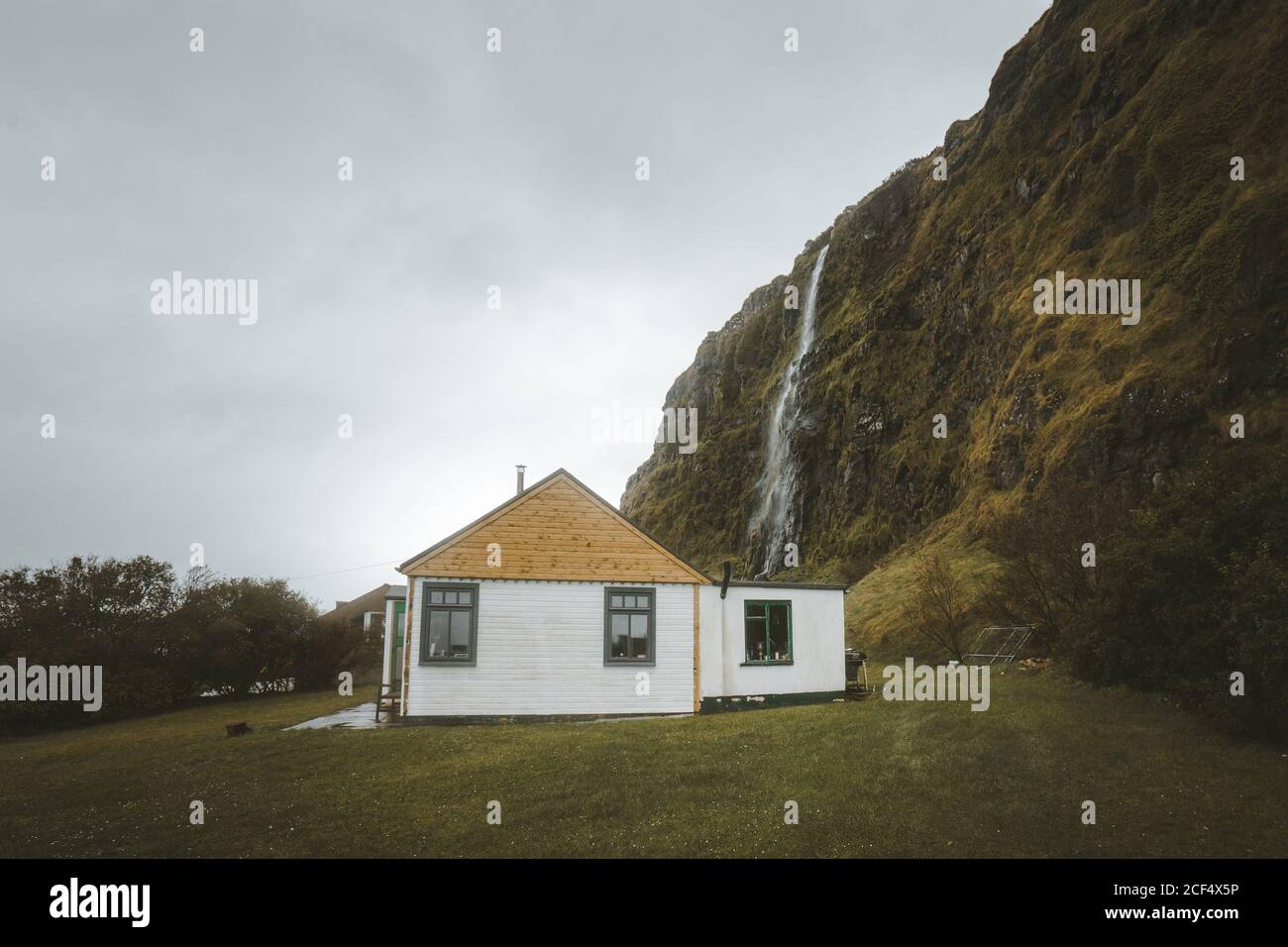 Kleines Landhaus aus Holz mit weißen Wänden und gelbem Giebeldach Dach auf grüner Wiese am Fuß der Klippe mit Wasserfall gegen grau bewölkten Himmel im Frühling Tag im Norden Irland Stockfoto