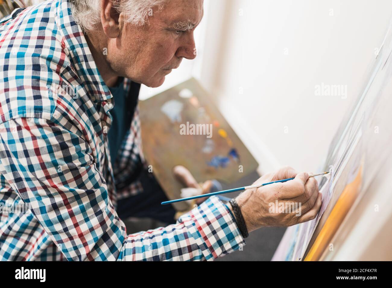 Seitenansicht von Crop Senior männlichen Künstler in legerer Kleidung Zeichnung Bild mit Pinsel auf Papier auf Staffelei halten Palette Mit Farben in der kreativen Kunstwerkstatt Stockfoto